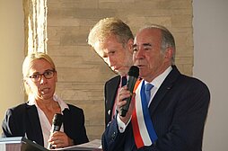 Bois-Colombes Bürgermeister Revillion spricht am Rednerpult ins Mikrophon, neben ihm stehen ein Mann mit Mikrophon und eine Frau s