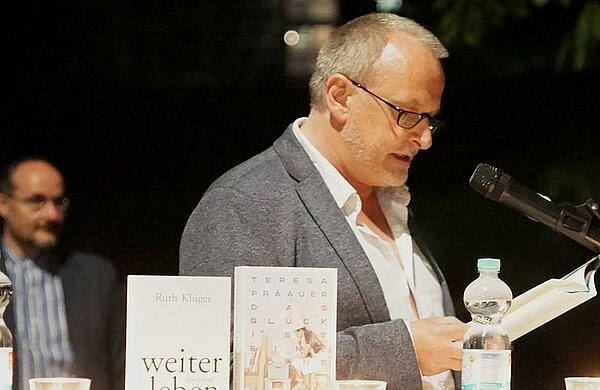 Ein Mann steht an einem Mikrofon und liest aus einem Buch