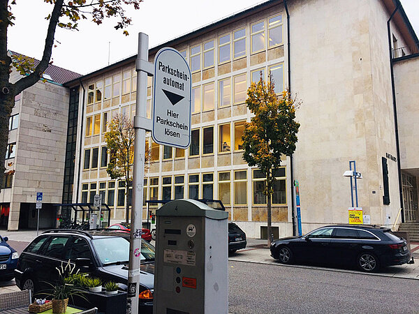 Ein Parkscheinautomat an einer Straße mit parkenden Autos, im Hintergrund das Neu-Ulmer Rathaus