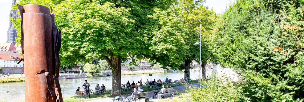 Menschen sitzen auf Stühlen und Bänken am Neu-Ulmer Donauufer