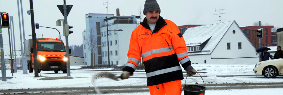 Ein Mann in orangener Schutzkleidung streut Splitt auf einen schneebedeckten Weg.
