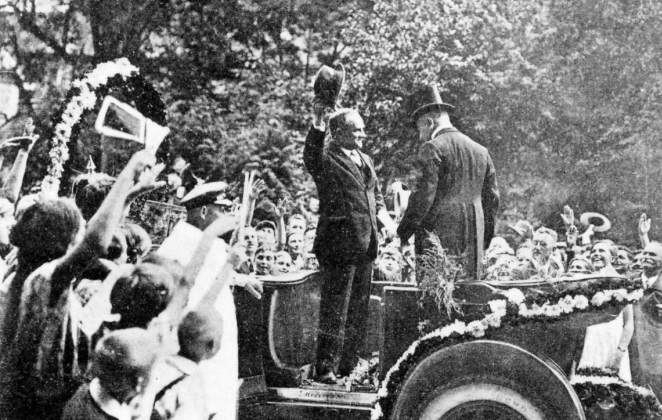 Schwarz-Weiß-Foto: Zwei Männer stehen in einem offenen Oldtimer, der eine Mann hebt seinen Hut und grüßt die jubelnde Menge, die um den Wagen steht.