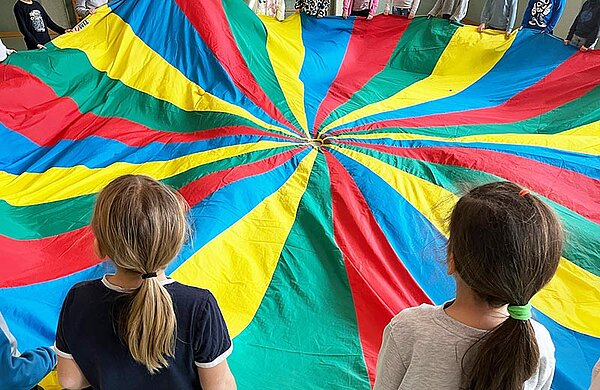 Kinder stehen im Kreis und halten ein Schwungtuch mit bunten Streifen.