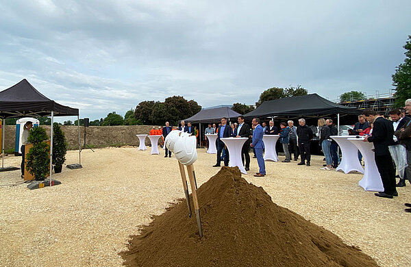 Mehrere Personen mit Baustellenhelmen stehen an einem Erdhaufen und werfen mit ihren Spaten Erde in die Luft.