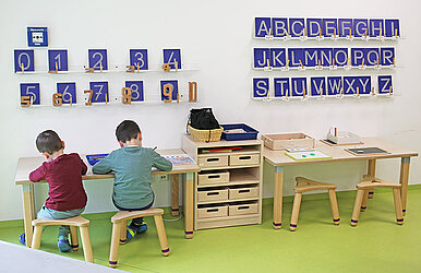 Zwei Kinder sitzen an einem kindergerechten Schreibtisch und malen, an der Wand vor ihnen sind das Alphabet und die Zahlen 0 bis 10 zu sehen.