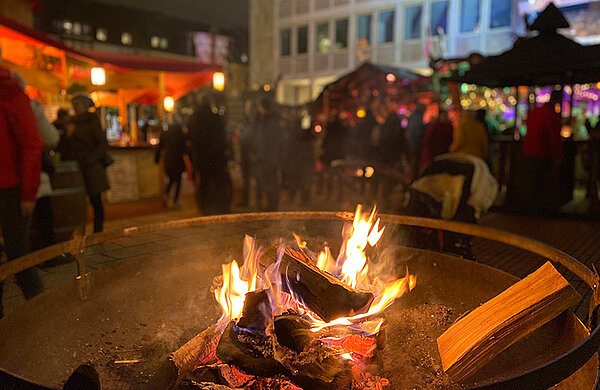 Feuerschale mit brennendem Holz, im Hintergrund Besucher des Weihnachtsmarktes