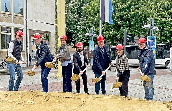 Mehrere Personen werfen beim symbolischen Spatenstich mit einem Spaten Sand in die Luft