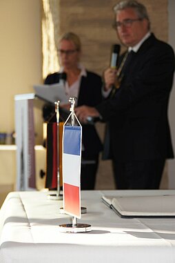 Im Hintergrund spricht OB Noerenberg am Rednerpult, im Vordergrund die Frankreichfahne als Aufsteller auf dem Tisch