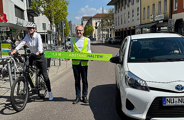 Oberbürgermeisterin Katrin Albsteiger auf dem Fahrrad am Fahrbahnrand. Sie wird von einem Auto überholt. Der Radverkehrsbeauftragte steht mit einem langen Maßband mit der Aufschrift "1,5 Meter Abstand halten" zwischen Fahrrad und Auto.