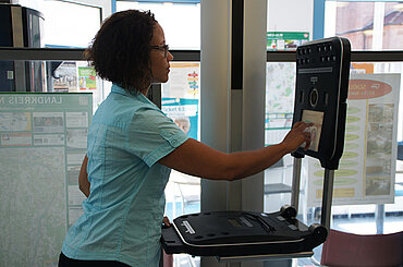 Foto von einer Frau während der Nutzung des Speed Capture-Terminals: Sie steht vor dem Gerät und tippt auf den Bildschirm, über dem eine Kamera integriert ist.