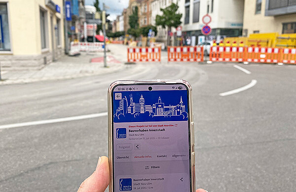 Smartphone mit Hinweis "Bauvorhaben Innenstadt" und der Neu-Ulmer Skyline, im Hintergrund eine Baustelle in der Neu-Ulmer Schützenstraße