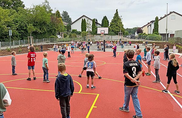 Schülerinnen und Schüler spielen auf dem neuen Platz mit Bällen, andere Mitschüler stehen und sitzen um den Platz und schauen zu.