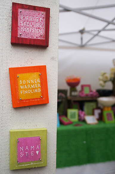 Stele mit drei quadratischen Kunstobjekten aus Holz und Glas, beschriftet mit den Begriffen "Eifriges Feuerknistern", "Sonnenwarmer Findling" und "Namaste", im Hintergrund ein Marktstand