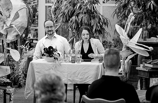 Julia Kulewatz und Florian L. Arnold sitzen an einem Tisch im Gewächshaus von Blumen Weimar.