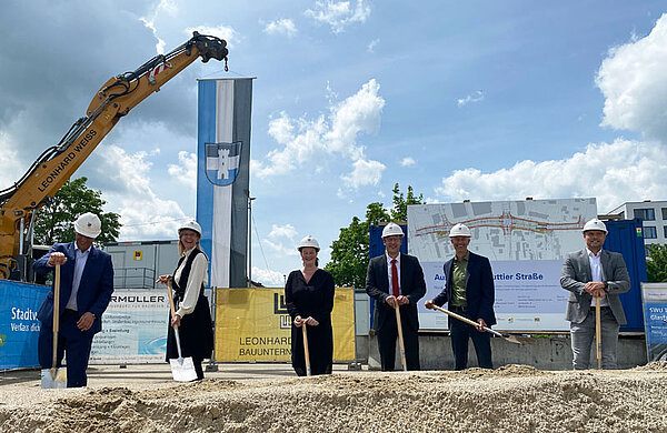 Oberbürgermeisterin Katrin Albsteiger und weitere Personen stehen nebeneinander auf einer Baustelle und stechen mit ihren Spaten in einen Kieshügel.