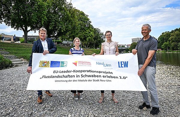 Die vier Personen stehen am Neu-Ulmer Donauufer und halten einen Banner mit der Aufschrift "EU-Leader-Kooperationsprojekt Flusslandschaften in Schwaben erleben 2.0"