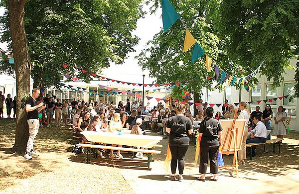 Sommerfest im Außenbereich der Kita, Kinder und Eltern sitzen an Tischen unter Bäumen