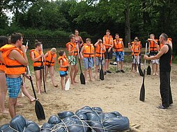 Die Jugendlichen tragen orangefarbene Rettungswesten und stehen mit Paddeln am Strand