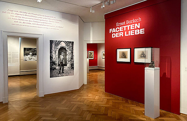 Blick in die Ausstellung "Ernst Barlach. Facetten der Liebe": Man sieht eine schwarz-weiß Fotografie des Künstlers an der Wand sowie drei seiner Werke vor einer roten Wand.