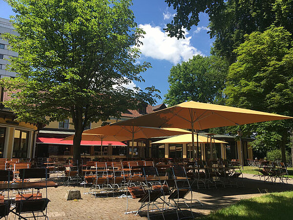 Biergarten mit Tischen, Stühlen und Sonnenschirmen unter Bäumen