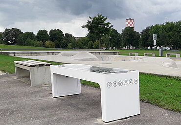 Eine weiße Sitzbank mit Piktogrammen an der Seite, im Hintergrund der Skatepool im Sport- und Freizeitpark Wiley
