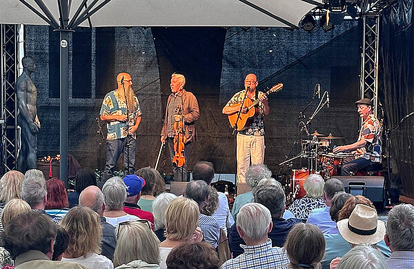 Eine Band spielt mit Geige, Gitarre und Schlagzeug auf einer Bühne vor Publikum.