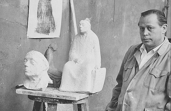 Gustav Seitz in seinem Außenatelier in Berlin-Pankow mit Gipsmodellen zum Kollwitz-Denkmal, 1958 (Foto: Gustav Seitz Stiftung) Schwarz-Weiß-Foto des Bildhauers neben zwei Gipsmodellen