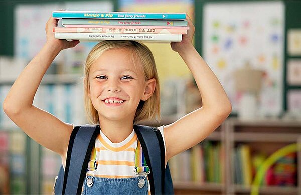 Ein Mädchen steht in einem Klassenzimmer und hält einen Stapel Bücher über den Kopf.