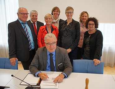 Oberbürgermeister Gerold Noerenberg sitzt an einem Tisch und unterzeichnet einen Vertrag, hinter ihm stehend 7 Personen