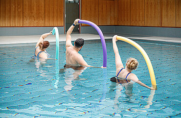 Drei Personen stehen in einem Schwimmbecken bis zum Bauch im Wasser und halten jeweils eine Schwimmnudel in die Höhe.