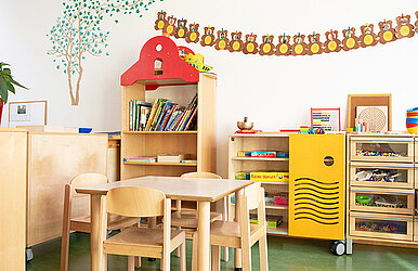  Kita-Gruppenraum mit Tisch und Stühlen sowie Regalen mit Büchern und Spielen