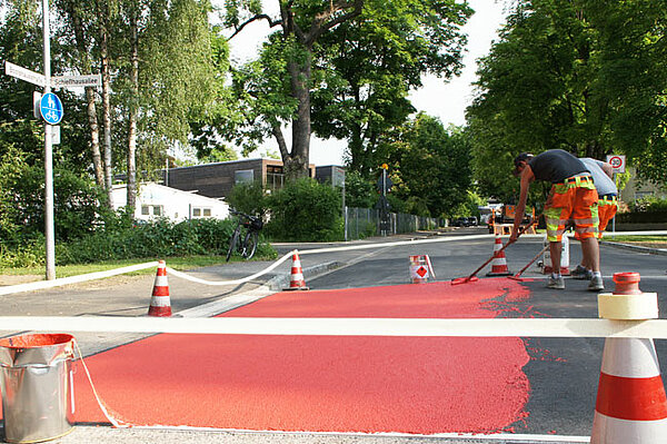 Mitarbeiter des Baubetriebshofes tragen im Kreuzungsbereich Schießhausallee/Bootshausstraße eine rote Farbmarkierung auf.