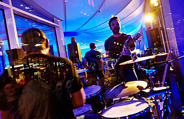 Blick durch die Scheibe in einen blau erleuchteten Raum, in dem eine Band mit Sänger, Drummer und Gitarrist auftritt