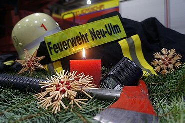 Tannenzweige und Strohsterne rund um eine brennende rote Kerze, im Hintergrund ein Feuerwehrhelm und ein Feuerwehranzug mit der  Aufschrift "Feuerwehr Neu-Ulm"