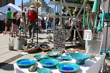 Im Vordergrund ein Marktstand mit bunten Keramikschälchen sowie weiteren Produkten, im Hintergrund schlendern Besucher über den Markt.