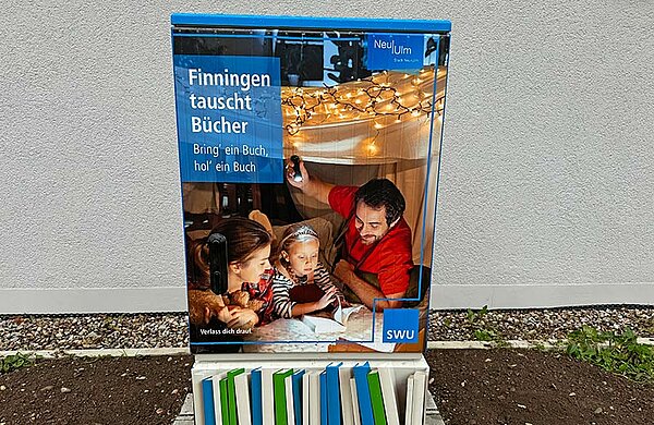 Bücherschrank mit der Aufschrift "Finningen tauscht Bücher" und der Abbildung von zwei Erwachsenen und einem Kind beim gemeinsamen Lesen