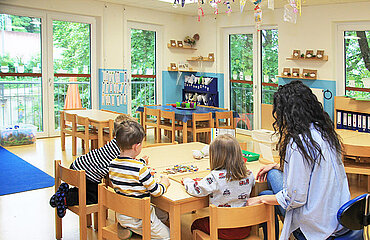 Drei Kinder und eine Betreuerin sitzen an einem Tisch im Gruppenraum der Kita.