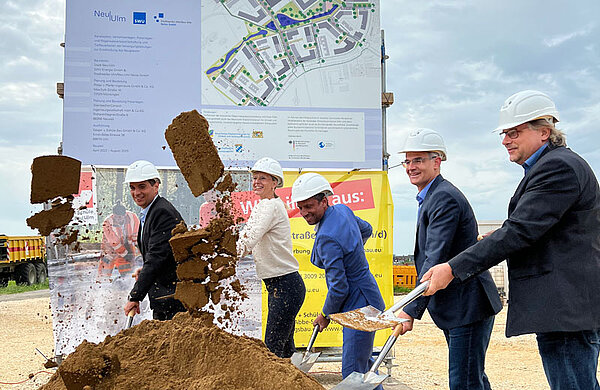Personen an Stehtischen bei einer Rede zum Spatenstich, im Vordergrund ein Erdhaufen mit Spaten.