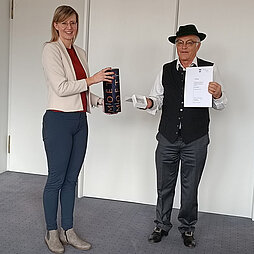 Horst Müller hält eine Urkunde sowie die Bürgermedaille in die Kamera, Oberbürgermeisterin Katrin Albsteiger überreicht ihm einen Geschenkkarton mit Champagner