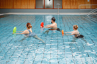 Drei Personen machen in einem Schwimmbecken eine Wassergymnastik-Übung mit einem Schwimmbrett