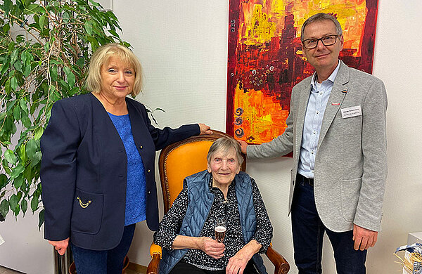 Die Jubilarin sitzt mit ein Glas Sekt auf einem Stuhl, rechts und links von ihr stehen Rosl Schäufele und Martin Pietschmann.