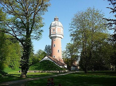 Das Bild zeigt den Kollmannspark: im Hintergrund das Wahrzeichen Neu-Ulms, der Wasserturm, im Vordergrund freistehende Bäume.