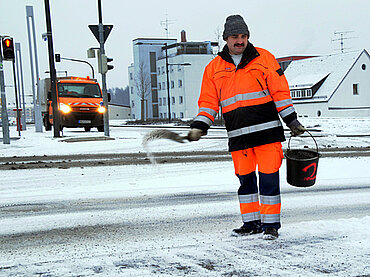 Ein Mitarbeiter des Baubetriebshofes wirft mit einer Schaufel Streugut auf die schneebedeckte Fahrbahn, in der anderen Hand hält er einen Eimer.