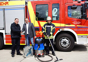 Drei Männer stehen vor einem Feuerwehrauto, zwei Männer geben sich die Hand. Der Brandmeister in Feuerwehrbekleidung hält den Schlauch des Wassersaugers in der Hand.