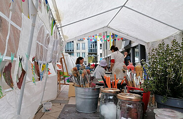 In einem Zelt hängen bunte Bilder und im Vordergrund stehen Behälter mit Stiften und Pinseln, im Hintergrund eine Gruppe mit Erwachsenen und Kindern in Malerkitteln
