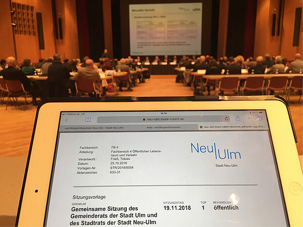 Im Vordergrund ein iPad mit der digitalen Sitzungsvorlage, im Hintergrund die Teilnehmer der gemeinsamen Sitzung