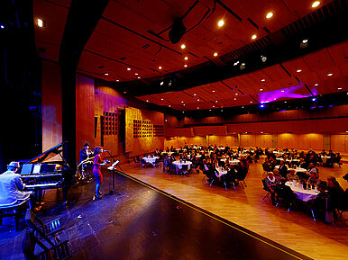 In einem großen Saal sitzen Menschen an gedeckten Tischen, auf der Bühne spielt eine Band.