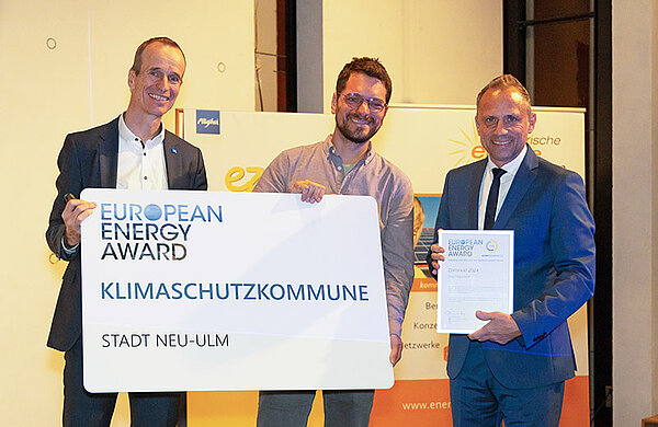 Drei Personen mit einem großen Schild mit der Aufschrift: European Energy Award Klimaschutzkommune Stadt Neu-Ulm