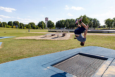 Ein Mann springt auf einem Trampolin in die Luft, im Hintergrund der Skatepark.