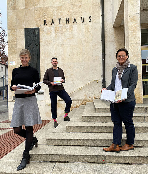 Oberbürgermeisterin Katrin Albsteiger, Dr. Larissa Ramscheid, die Leiterin des Stadtarchivs und Autor Eberhard A. Merk mit jeweils einer Ortschronik in der Hand vor dem Neu-Ulmer Rathaus